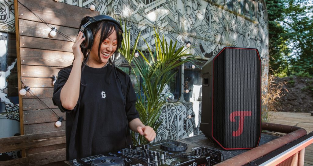 Frau steht im Freien am DJ-Mischpult, trägt dabei DJ-Kopfhörer, daneben steht der ROCKSTER AIR 2