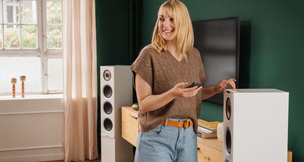 STEREO L 2 Lächelnde Frau mit Smartphone steht neben einem weißen Lautsprecher